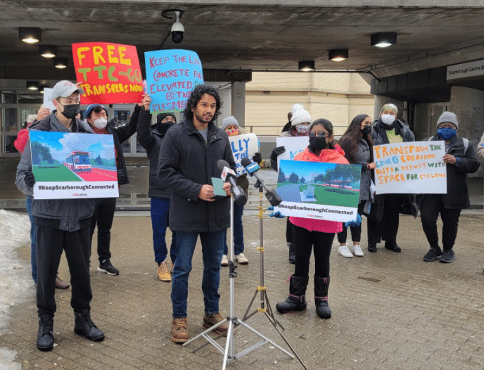 A group of people holdinh up several colourful signs as they stand outside in front of a subway street entrance.