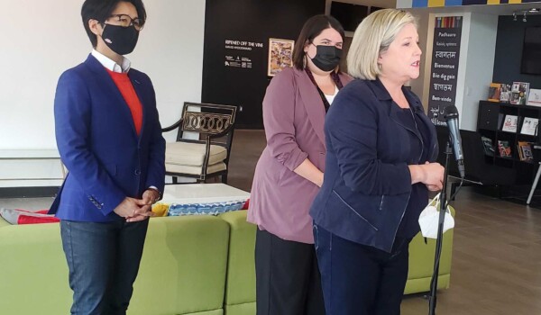 Three people standing in front of a green couch as they speak to a microphone. One person has a blue blazer, the person in the middle has a lavender coat and the person on the far right wears a navy jackey and has blonde hair.