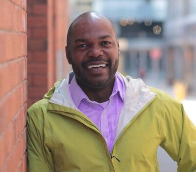 A man wearing a lavender dress shirt with a mustard jacket on top. He is smiling and leaning on a brick wall.