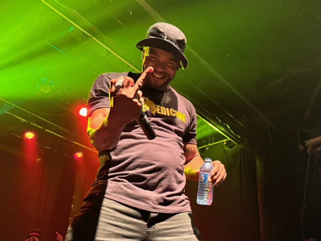 Method Man, standing onstage in a bucket hat, holds a water bottle in his left hand while holding a microphone and flipping off the camera with his right hand