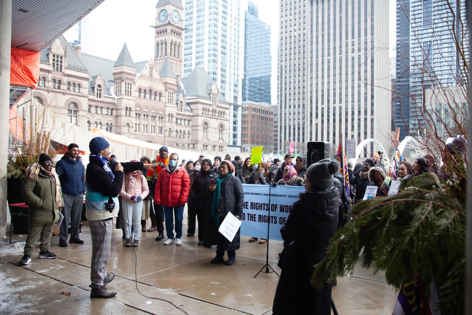 A crowd of people holding banners and signs in front of city hall. A speaker using a mic faces the ralliers.