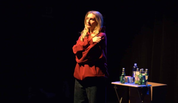 Actress dressed in black jeans, a red hoodie, with a concerned or somewhat exasperated look on her face, in a self hug sort of posture. She is lit by stage light, standing next to a small folding table with 4 small multicolored plastic tumbler glasses, a 6-pack of Peroni beer bottles, and a water bottle. The background is dark, making her a strong focal point amidst the black.
