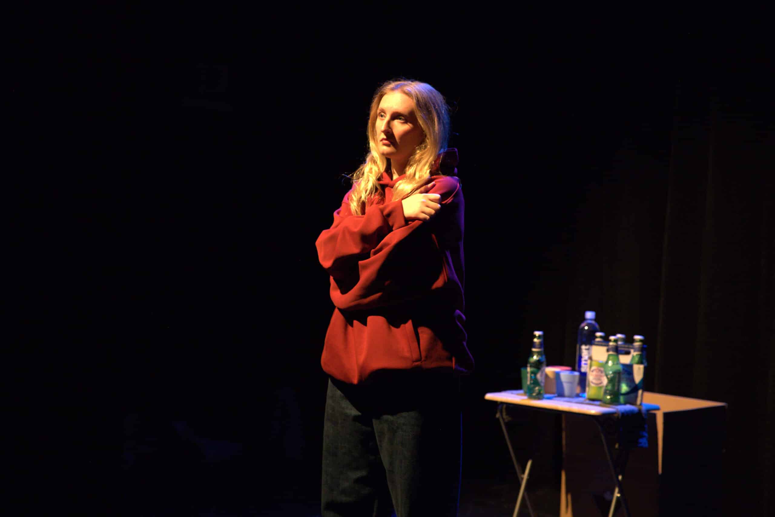 Actress dressed in black jeans, a red hoodie, with a concerned or somewhat exasperated look on her face, in a self hug sort of posture. She is lit by stage light, standing next to a small folding table with 4 small multicolored plastic tumbler glasses, a 6-pack of Peroni beer bottles, and a water bottle. The background is dark, making her a strong focal point amidst the black.