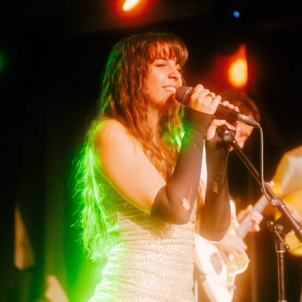 Ella Forrest, in a shimmery silver outfit, with long black fingerless gloves, singing on stage while holding a microphone resting in a stand. A stagelight behind her is illuminating her hair a bright green.