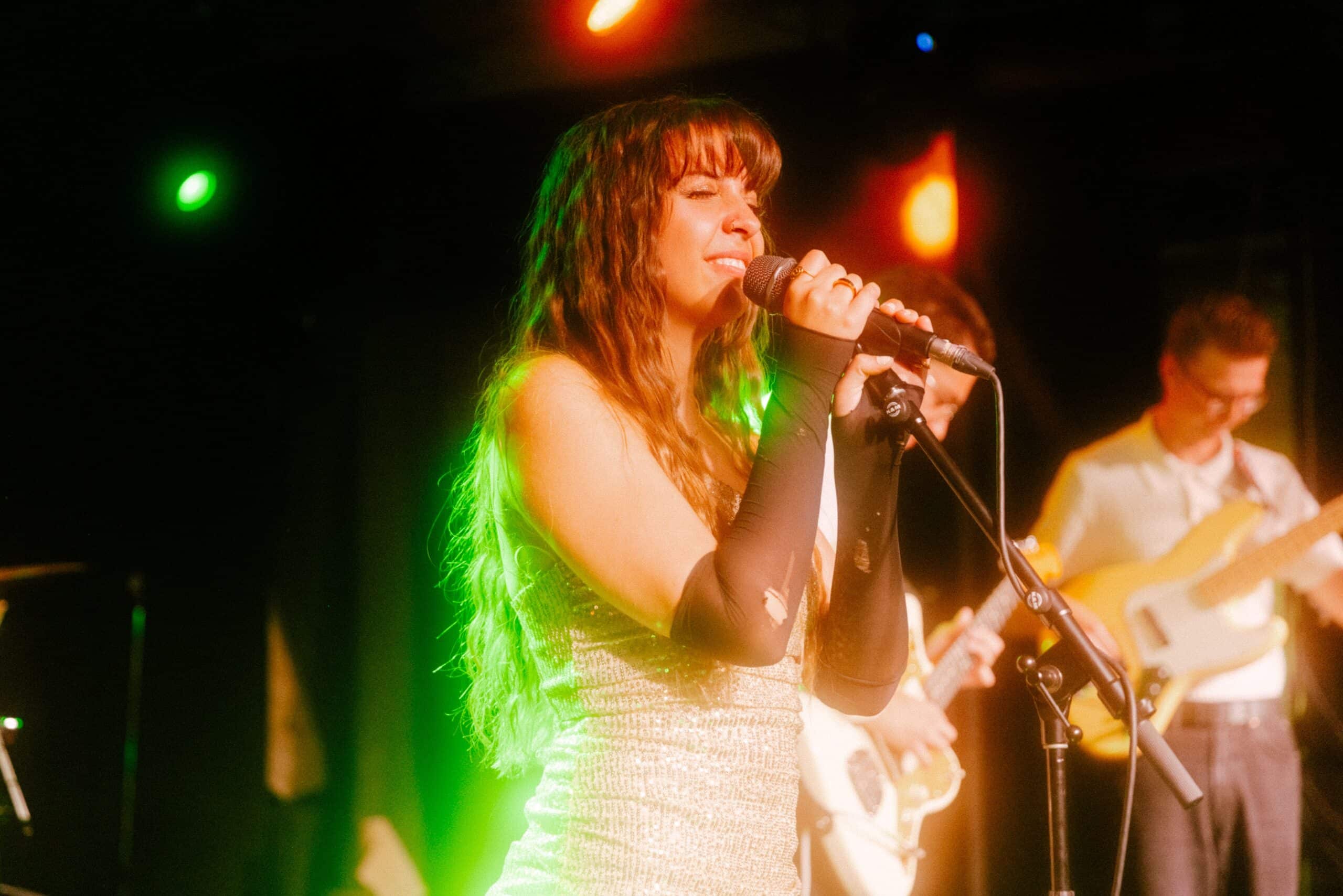 Ella Forrest, in a shimmery silver outfit, with long black fingerless gloves, singing on stage while holding a microphone resting in a stand. A stagelight behind her is illuminating her hair a bright green.