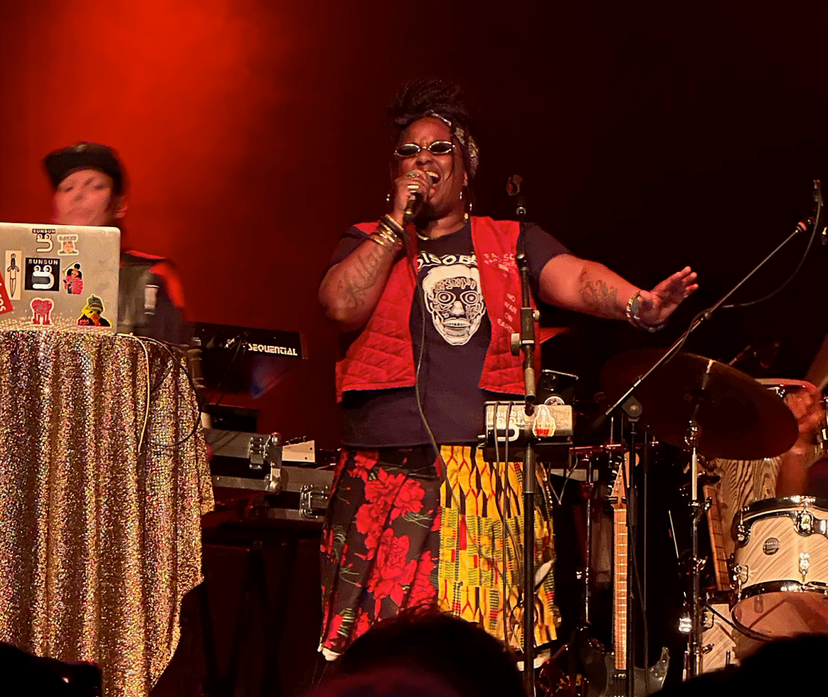 Witch Prophet singing into a mic centre stage with other arm outstretched over the audience, DJ in the background with a Macbook laptop covered in stickers.
