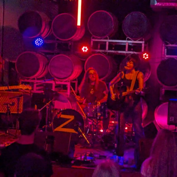Zoh Amba in bluejeans and a black tank top plays an acoustic guitar, mouth open as she sings into a mic, with electric guitarist hanging his head down and his shaggy hair covering his face. A drummer in the background with long hair and square framed glasses plays along. The stage is in blue and red lights with the round tops of beer kegs lined in rows behind them as the stage backdrop.